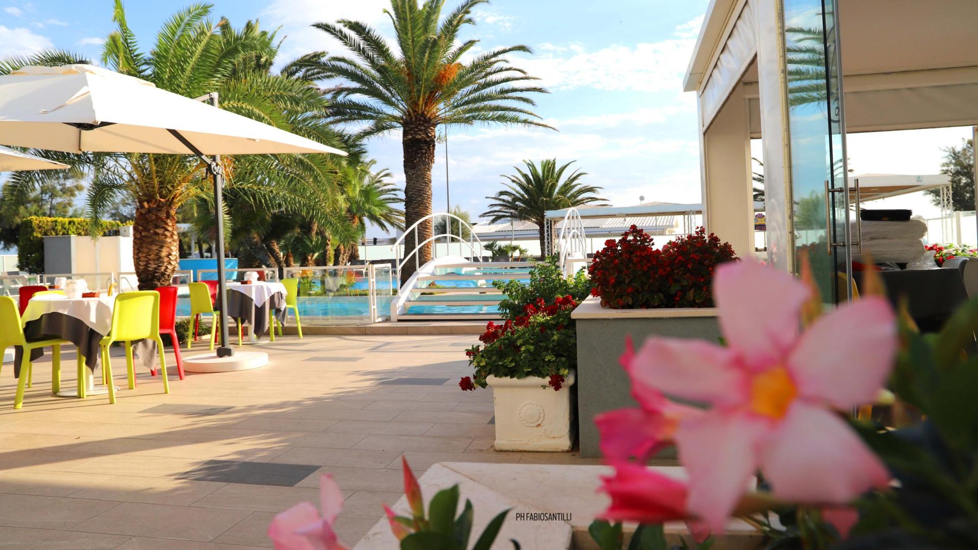 Terrazza con tavoli colorati, palme e piscina, atmosfera estiva e rilassante.