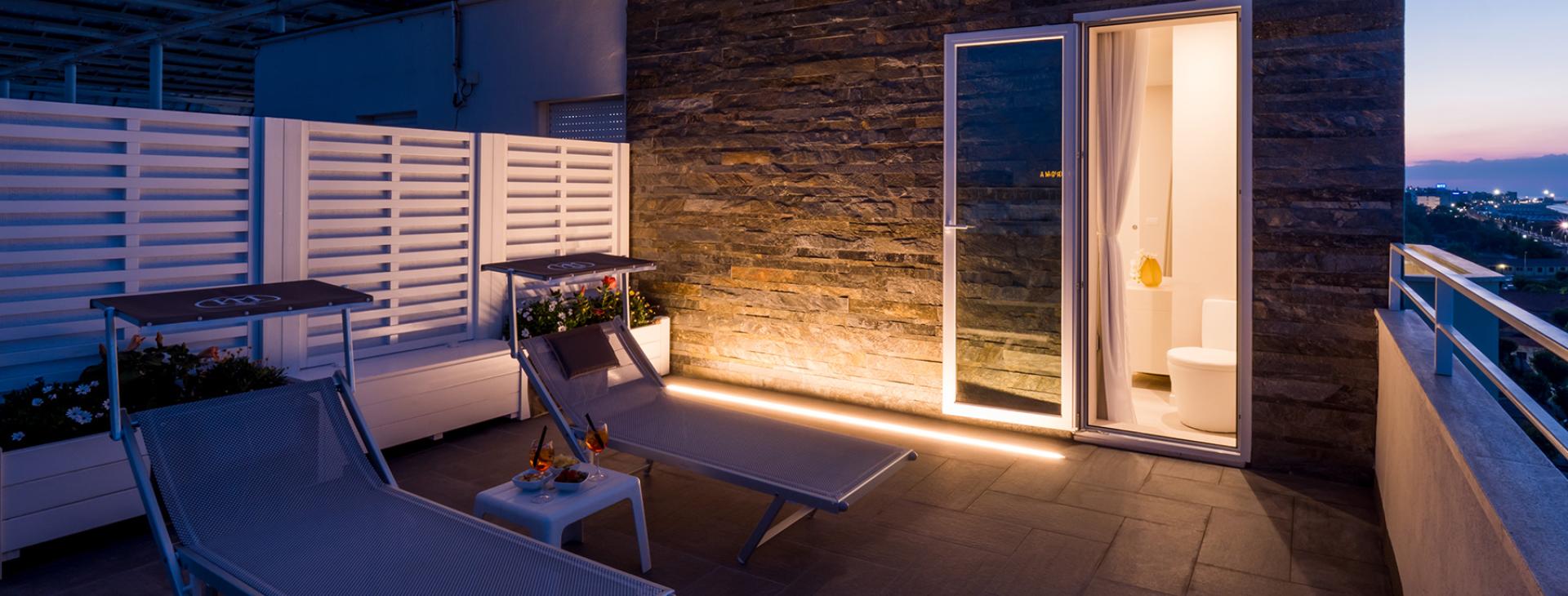 Terrazza con lettini, tavolino e vista notturna, accesso a bagno illuminato.