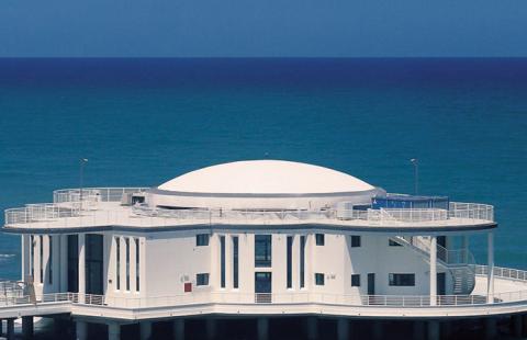 Edificio circolare su palafitte, affacciato sul mare con cielo azzurro.