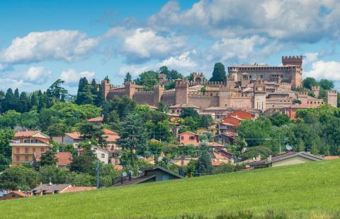 Castello medievale circondato da case e verdi colline, sotto un cielo azzurro.