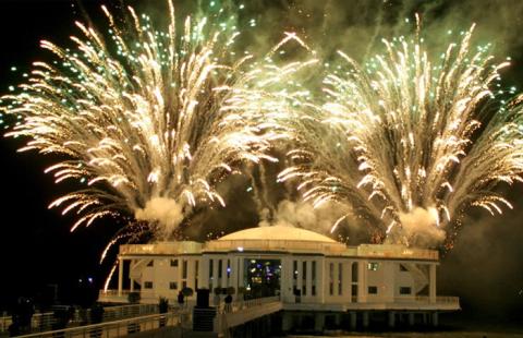 Fuochi d'artificio illuminano una struttura sul mare di notte.