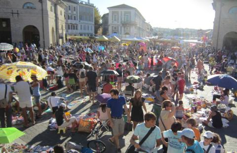 Folla in una piazza con ombrelloni e bancarelle durante un mercato all'aperto.