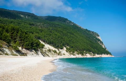 Spiaggia con sabbia bianca, mare turchese e colline verdi sullo sfondo.