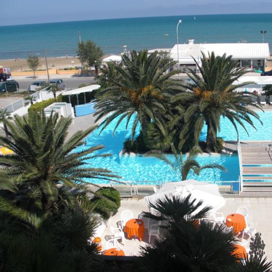 Piscina con palme vicino alla spiaggia, vista mare e sedie arancioni.