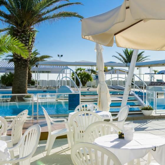 Piscina all'aperto con sedie bianche e ombrelloni, circondata da palme e fiori.