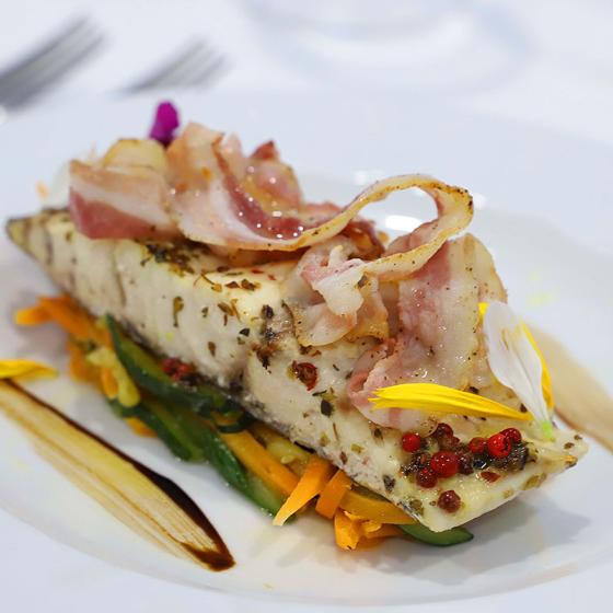 Filetto di pesce con pancetta, verdure e decorazioni floreali su piatto elegante.