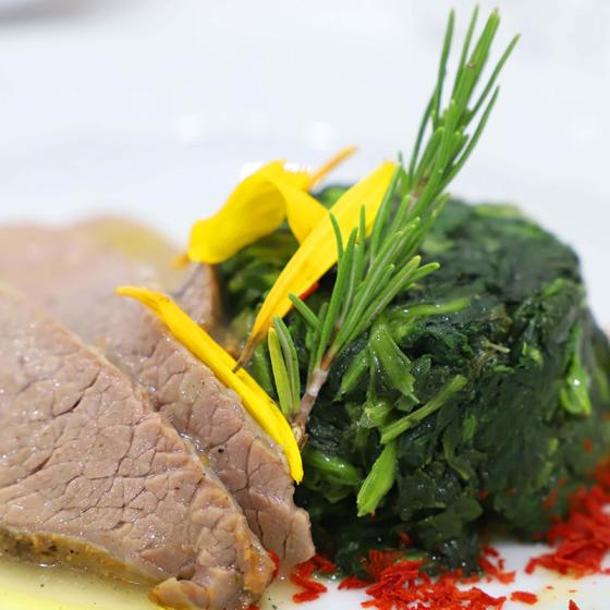 Carne con spinaci, decorata con petali gialli e rosmarino su piatto elegante.