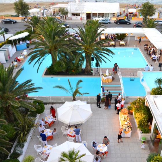 Piscina all'aperto con palme, tavoli apparecchiati e vista sulla spiaggia.