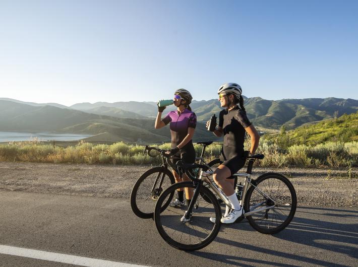 Due ciclisti si fermano a bere, ammirando il paesaggio montano al tramonto.