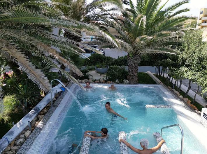 Piscina all'aperto con persone che si rilassano tra palme e vegetazione.